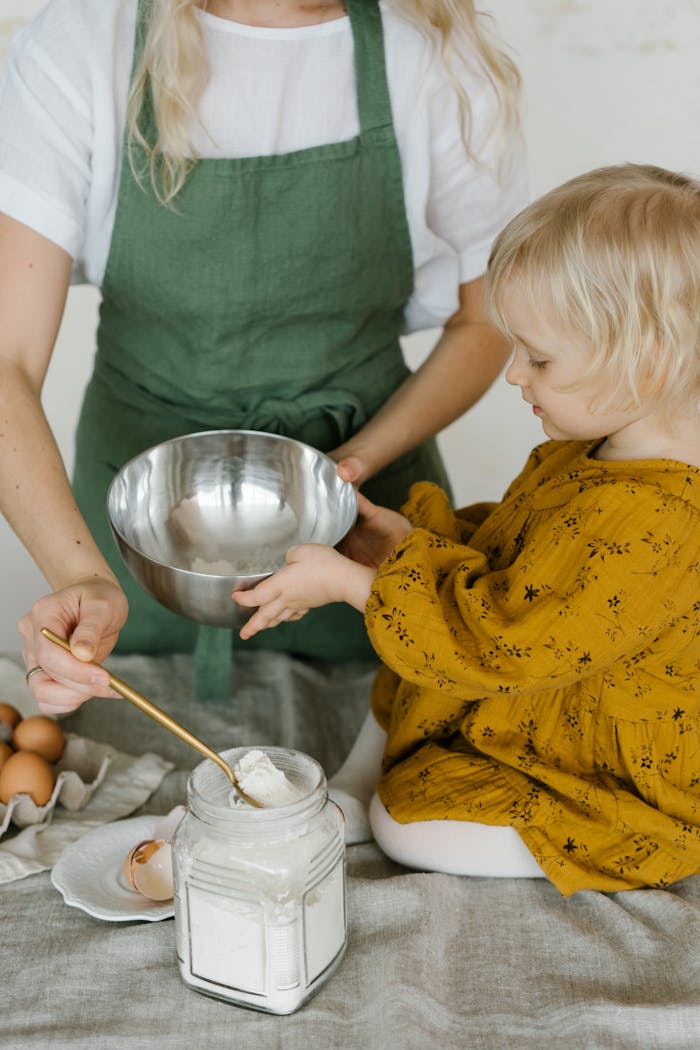 High angle of little daughter sitting on table with stainless bowl helping mother in making dough